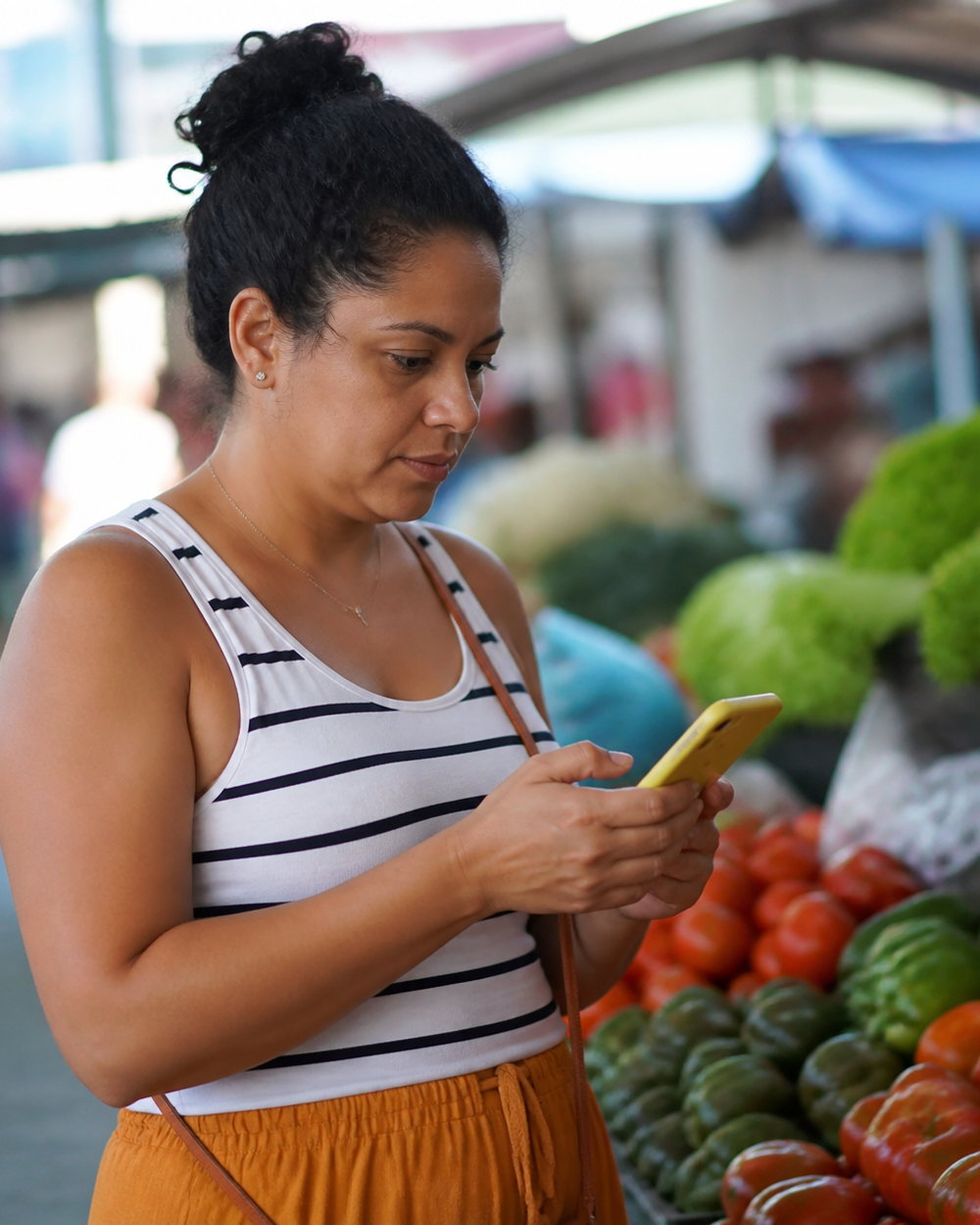 Consumidora consultando o celular em frente a uma banca de frutas e verduras em feira livre.